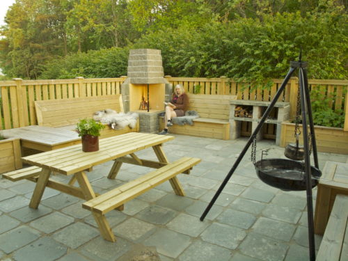 Austrått agrotourismus, Kårstua, schiefer als terassenbelag, feuerpfanne auf der rechten seite, esstisch mit bänken auf der linken seite, im hintergrund liest eine frau ein buch neben einem Kamin